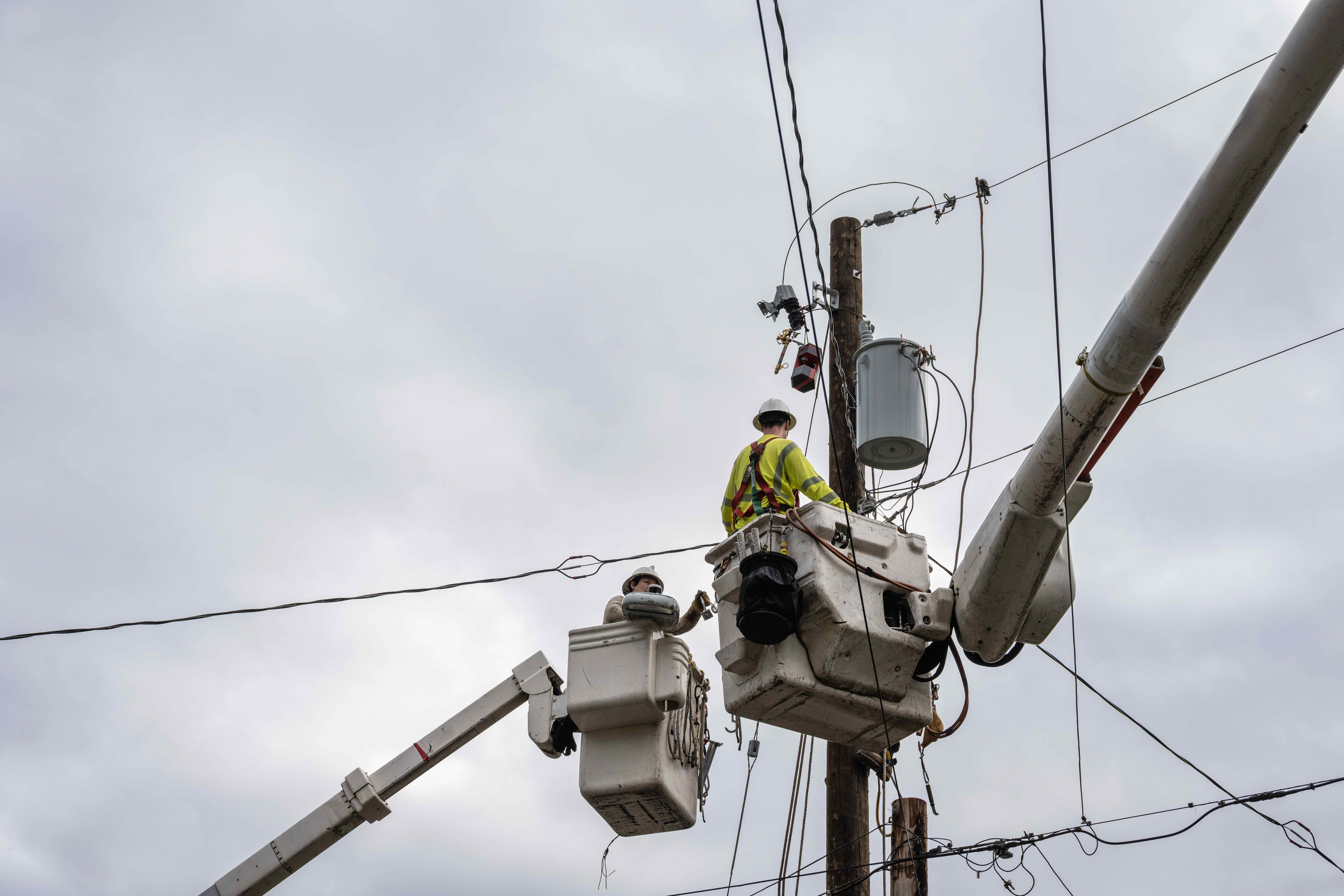 Utility workers in bucket trucks repairing electrical equipment on a power pole with overhead lines on a cloudy day.